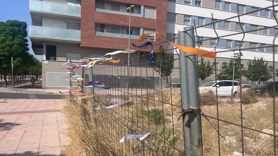Valla de la parcela S-9 cubierta de lazos de colores con los deseos del barrio, y los bloques de viviendas al fondo